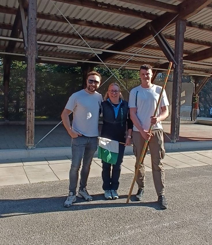 Three individuals pose under a gazebo. A man on the left, a woman in the middle, and a man on the right holding a flag.