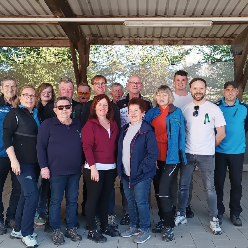 A group of people wearing various jackets and sneakers poses for a photo under a pavilion with a metal roof and glass walls. Trees are visible in the background.