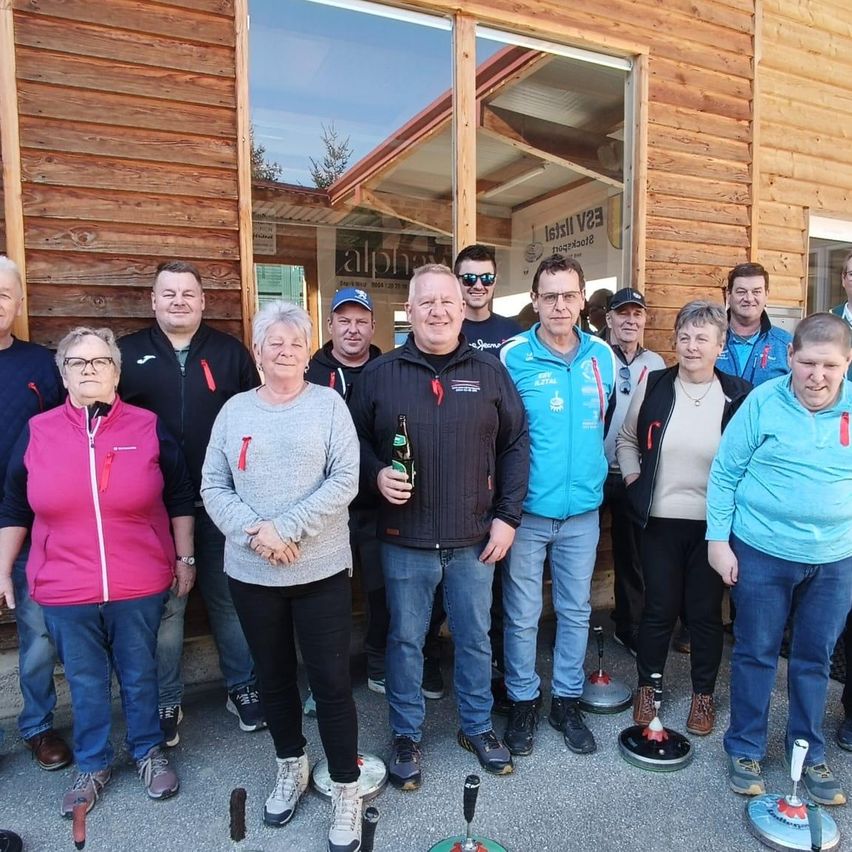 A group of people pose for a photo outside a wooden building. They are holding curling brooms and wearing winter clothing.