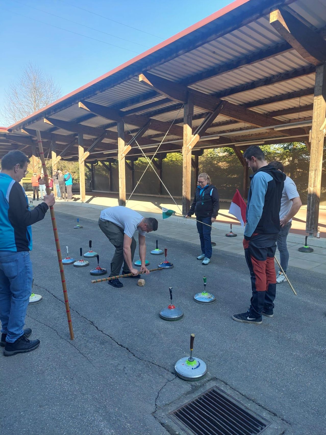 A group of people are playing a game with mallets and balls on a paved surface under a wooden structure.