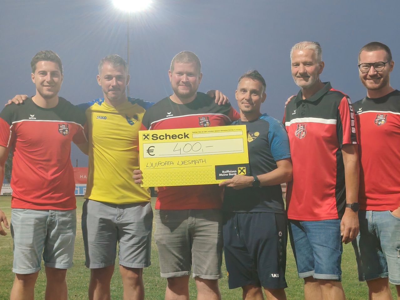 Fünf Männer stehen auf einem Rasenfeld, einer hält einen gelben Scheck über 400 Euro. Sie sind in Sporttrikots und Shorts gekleidet. Hinter ihnen steht ein Laternenpfosten und ein klarer Himmel.