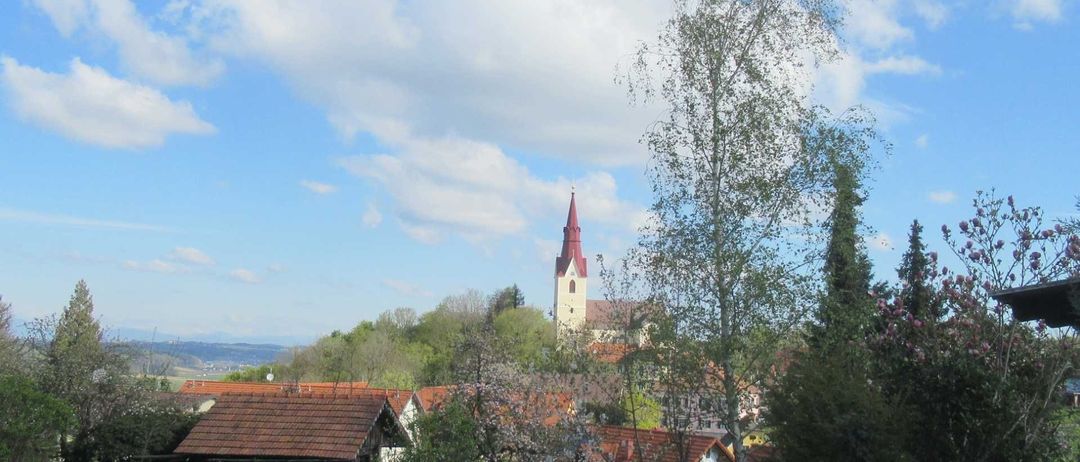 Ein Blick auf eine Stadt mit einem Kirchturm in der Mitte, umgeben von Bäumen und Häusern mit roten Dächern. Der Himmel ist blau mit einigen Wolken.