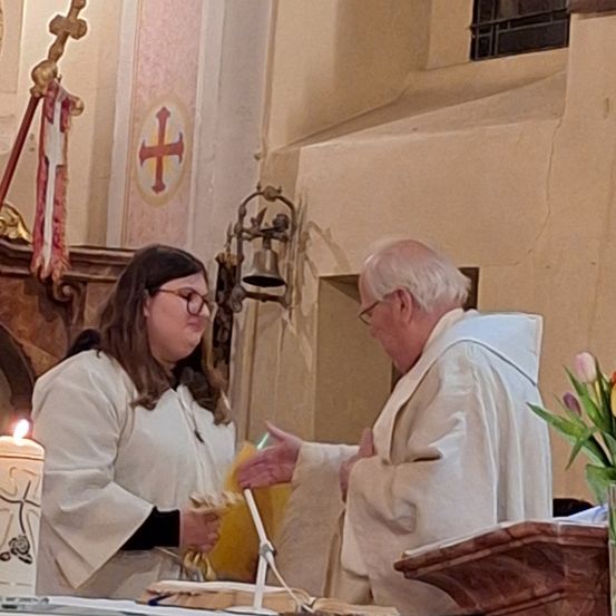 Zwei Personen, eine Frau und ein älterer Mann, sind in einer Kirche in weiße Kutten gekleidet. Die Frau hält eine Kerze und etwas gelbes Gewebe, während der Mann mit einer Geste und einem gelben Tuch in der Hand steht. Eine Glocke hängt an der Wand, und ein Kreuz ist sichtbar.