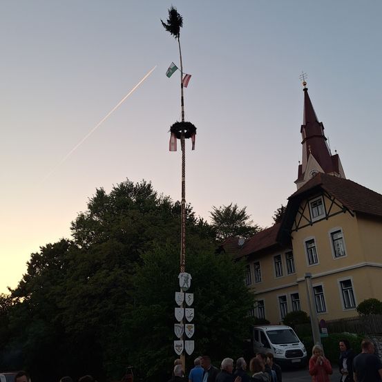 Ein hoher Mast mit mehreren Flaggen und Wappen steht vor einer Kirche. Es haben sich Menschen darum versammelt, und ein weißer Van ist in der Nähe geparkt. Der Himmel ist klar, und die Sonne geht unter.