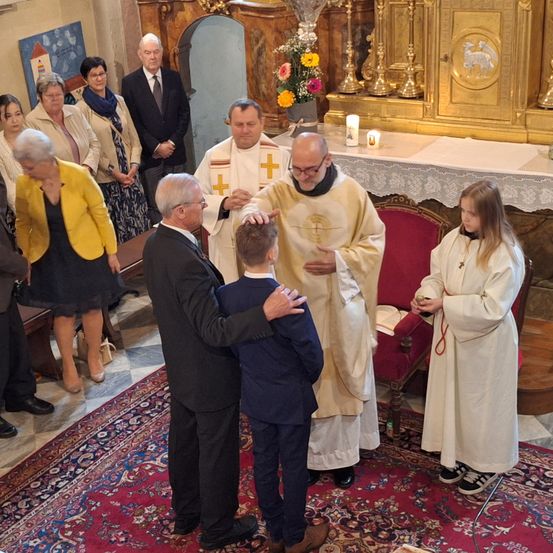 Ein Priester segnet einen jungen Jungen während einer Zeremonie in einer Kirche. Ein erwachsener Mann hält den Jungen an der Schulter. Ein Mädchen steht in der Nähe und trägt ein weißes Gewand.