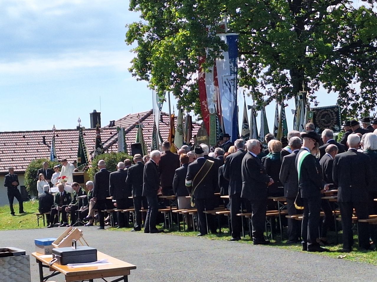 A group of people in suits standing under a tree. Flags are on poles behind them. A man in a white robe stands in front of a table.