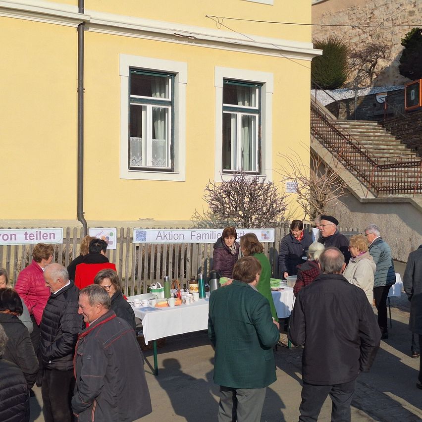 A group of elderly people are standing around a table with food, drinks, and cups. They are in front of a yellow building with windows and a sign that reads 'Aktion Familie'.