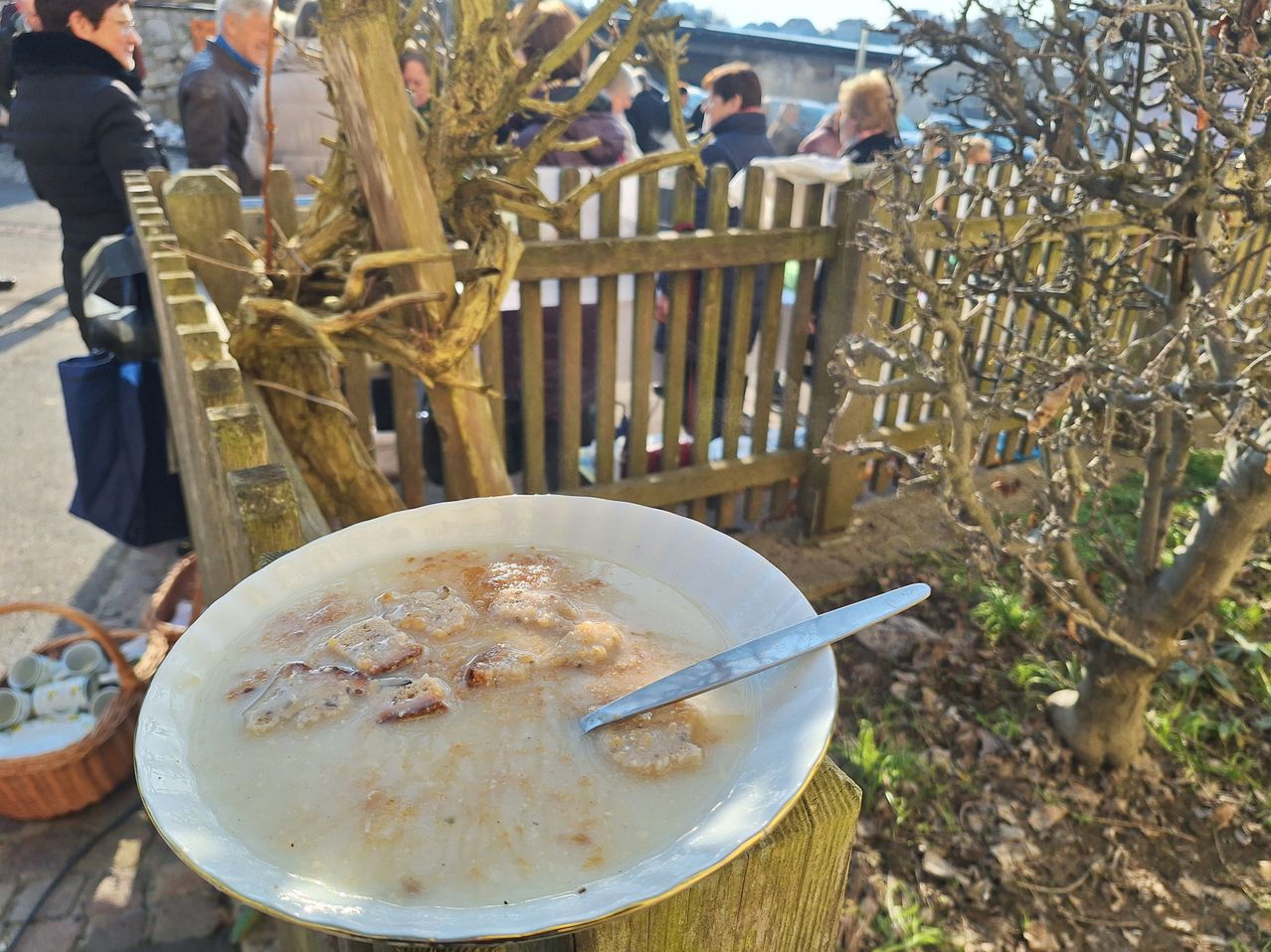 A bowl of soup with a spoon sits on a wooden post in an outdoor setting, with people standing behind a fence in the background.