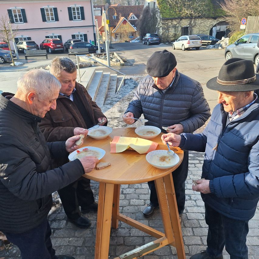 Four elderly men gather around a small wooden table on a cobblestone street. Each holds a plate with food, and they appear to be sharing a meal. Behind them, a building with many windows stands, and several cars are parked.