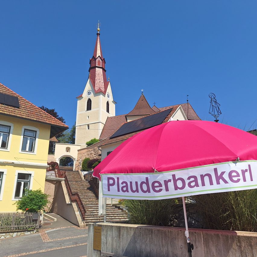 Ein leuchtend rosafarbener Regenschirm mit dem Wort Plauderbankerl darauf steht vor einer Kirche mit einem Turm und Solarpanelen auf dem Dach.