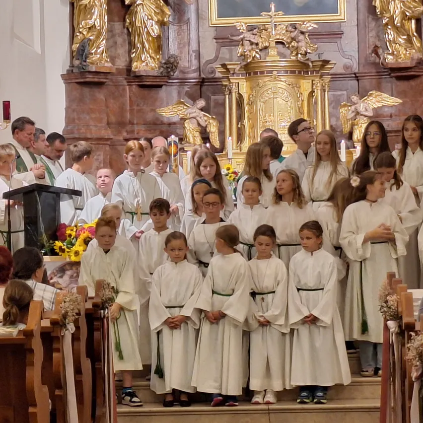 Eine Gruppe von Kindern in weißen Gewändern steht in einer Kirche, wahrscheinlich während einer religiösen Zeremonie. Sie stehen vor einem Altar mit einer goldenen Statue und einem Kreuz.