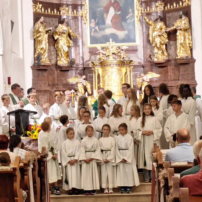 Ein Chor von Kindern in weißen Gewändern steht vor einem Altar in einer Kirche, mit goldenen Statuen und einem großen Gemälde dahinter.