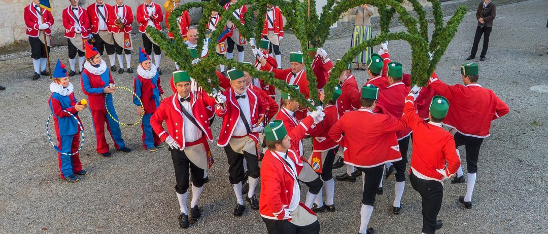 Eine Gruppe von Menschen in roten und weißen Uniformen und grünen Hüten hält einen großen Kranz aus immergrünen Pflanzen. Sie führen eine Aufführung im Freien durch. Hinter ihnen steht ein Gebäude mit einem Kreuz oben.