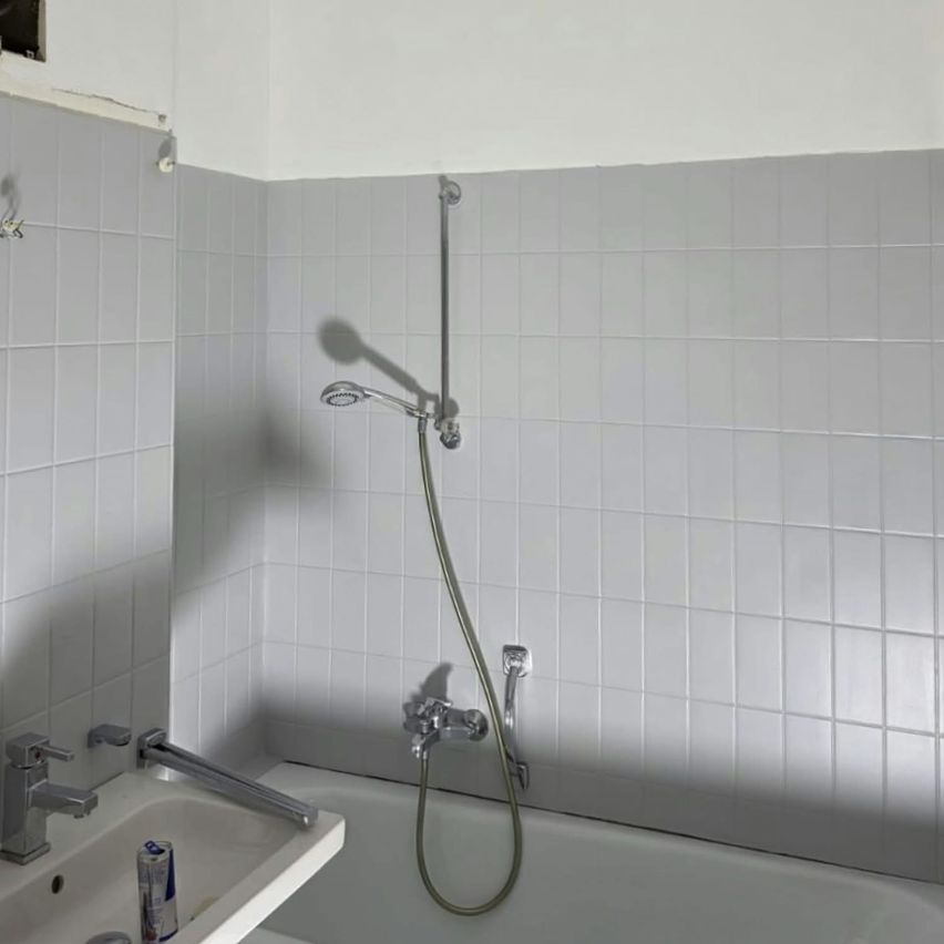 A bathroom with white tiles on the walls and a bathtub. A showerhead and faucet are attached to the wall above the bathtub. A can is placed on the sink.