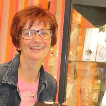 Eine Frau mit roten Haaren, die eine Brille und ein rosa T-Shirt trägt, steht vor einer Schmuckvitrine. Sie hält eine schwarze Schachtel mit einem goldenen Adleranhänger.