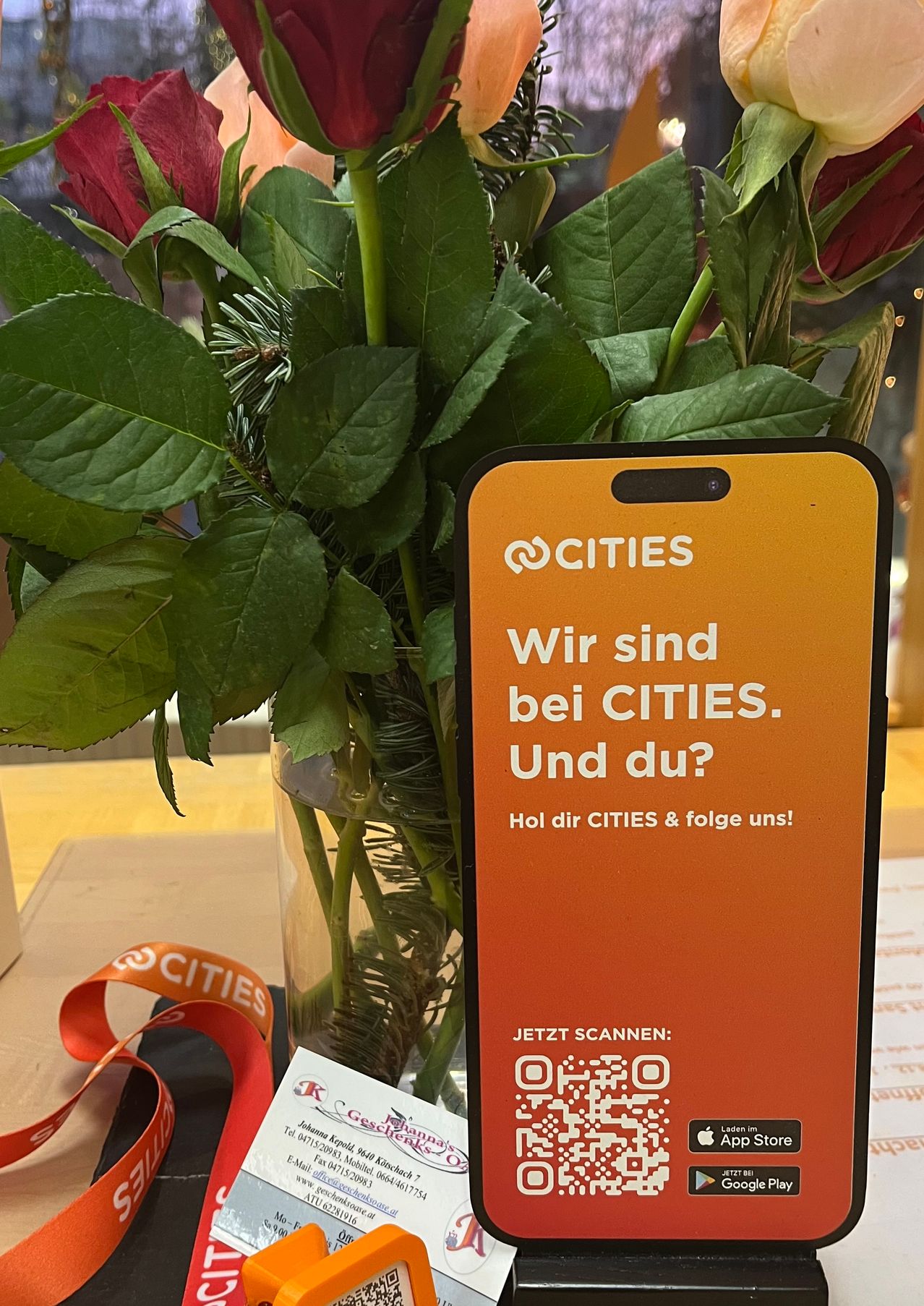 Ein orangefarbener Smartphone-Bildschirm zeigt den Text 'Wir sind bei CITIES. Und du?' mit einem QR-Code darunter. Ein Strauß roter Rosen steht in einem Glasvase hinter dem Handy.