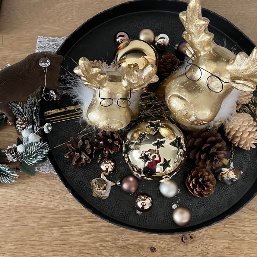 Eine schwarze Schale enthält goldene Rentier-Ornamente, verschiedene goldene und silberne Weihnachtskugeln und Tannenzapfen. Die Schale steht auf einem Holztisch, mit zusätzlichen Weihnachtsdekorationen darum herum.