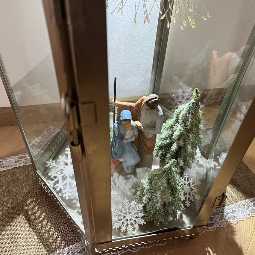 Eine Glaslaterne mit Krippenfiguren, einem Weihnachtsbaum und Schneeflocken im Inneren. Sie steht auf einem Holztisch mit Spitzenbordüre.