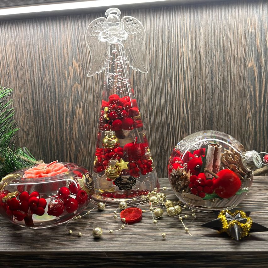 Drei Glas-Weihnachtsschmuckstücke stehen auf einem Holztisch. Das höchste zeigt ein Engeldesign mit roten Beeren und goldenen Sternen. Die anderen enthalten rote Blumen und Zimtstangen.