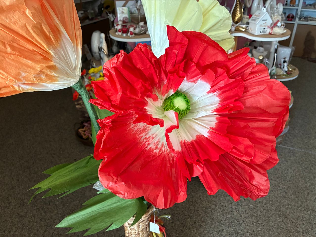 Eine leuchtend rote Papierblume mit einem weißen Zentrum und grünem Stiel ist in einem Geschäft mit anderen Artikeln.