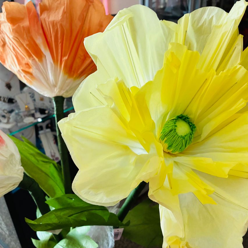 Eine leuchtend gelbe Papierblume mit grünen Stempeln in der Mitte, umgeben von anderen Papierblumen, wird auf einem Tisch ausgestellt.