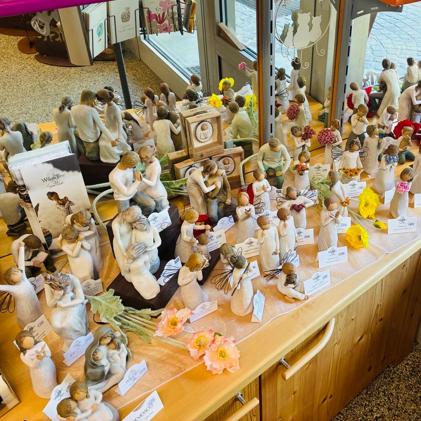 Ein Geschäft zeigt eine Vielzahl von Figuren, jede mit einzigartigen Posen und Blumen, auf einem Holztisch angeordnet. Einige Figuren haben Preisschilder. Die Ausstellung spiegelt sich in einem Glasfenster wider.