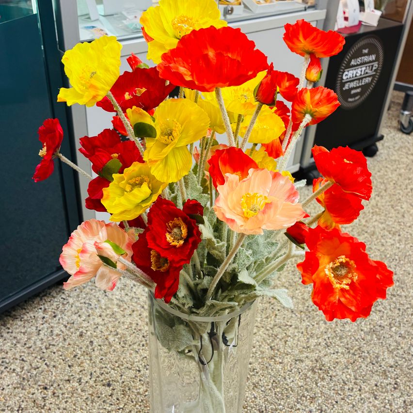 Ein Glasvase mit verschiedenen leuchtend gefärbten Blumen, darunter rot, gelb und orange, steht auf einem strukturierten Boden.