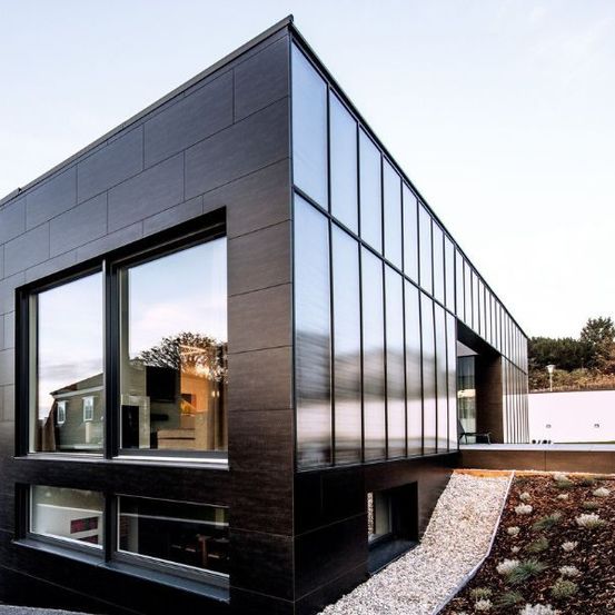 Ein modernes Haus mit schwarzer Fassade und großen Fenstern, die den Himmel und die Bäume reflektieren. Das Haus ist von einem Garten mit Kies und Pflanzen umgeben.