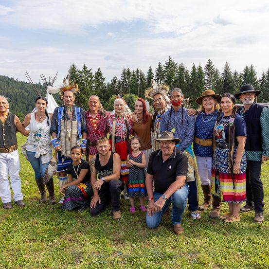 Eine Gruppe von Menschen, einschließlich Erwachsener und Kinder, posiert für ein Foto auf einem Grasfeld mit Bäumen und Tipis im Hintergrund.
