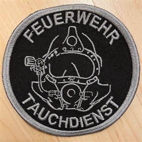 Ein schwarzer kreisförmiger Aufnäher mit Taucherhelm und der Aufschrift 'Feuerwehr Tauchdienst'. Er befindet sich auf einer Holzoberfläche.