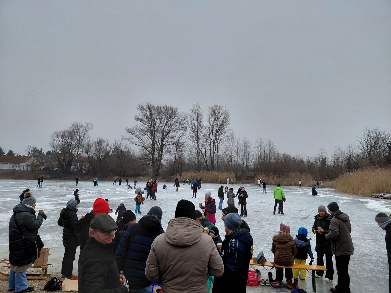 Eine Gruppe von Menschen, die auf einem zugefrorenen See Schlittschuh laufen, einige tragen Mützen und Brillen. Zuschauer stehen an der Seite und beobachten. Bäume umgeben das Gebiet, und der Himmel ist bedeckt.