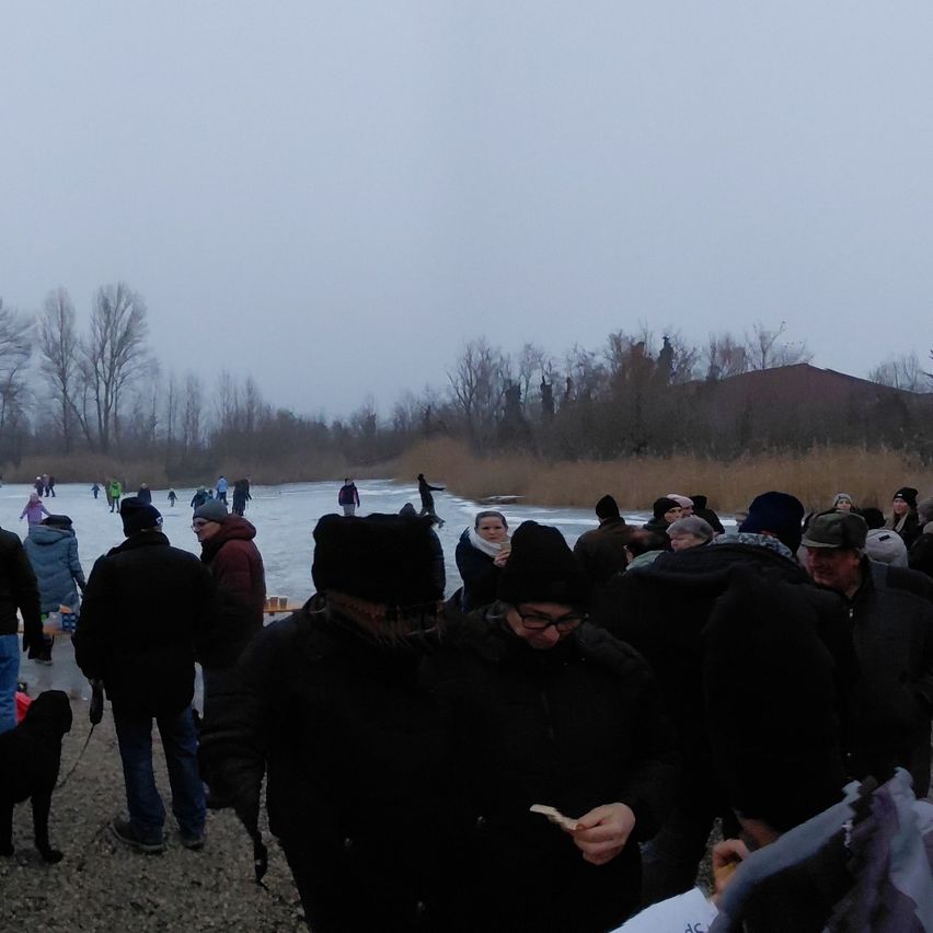 Eine Menschenmenge versammelt sich in der Nähe eines vereisten Sees, viele tragen Winterkleidung. Einige skats auf dem Eis, während andere am Ufer stehen. Ein Gebäude ist im Hintergrund zu sehen.