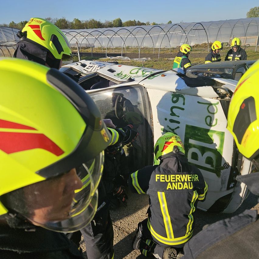 Feuerwehrleute in gelben Helmen retten eine Person aus einem beschädigten Fahrzeug. Der Schauplatz ist im Freien mit Gewächshäusern im Hintergrund.