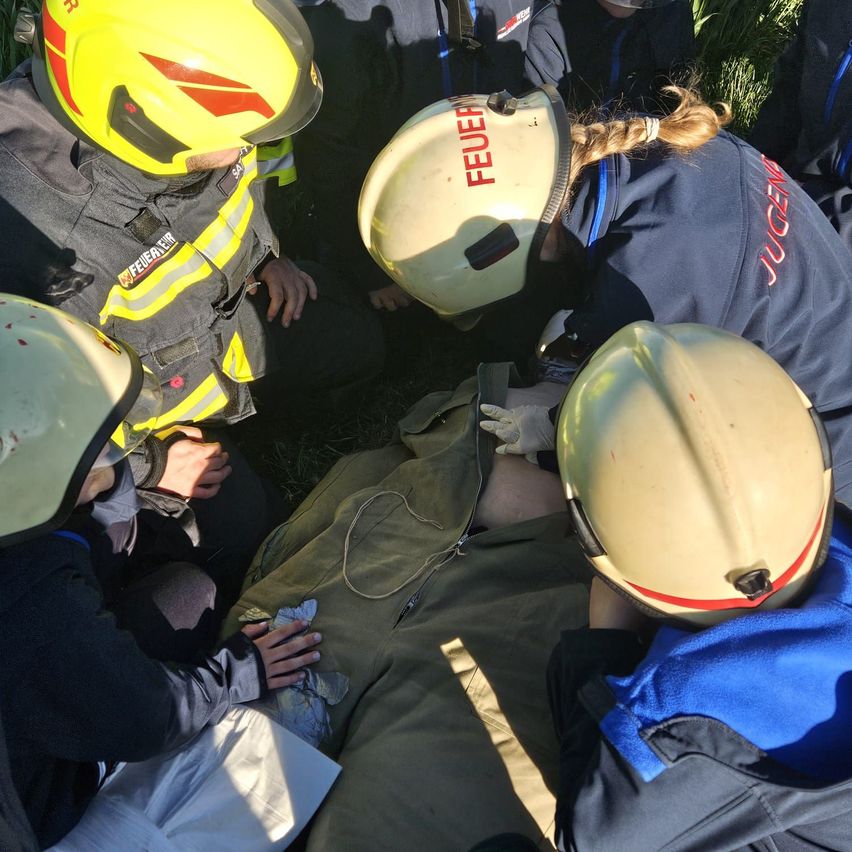 Eine Gruppe von Feuerwehrleuten mit Helmen führt eine Rettungsaktion an einer auf dem Boden liegenden Person durch.
