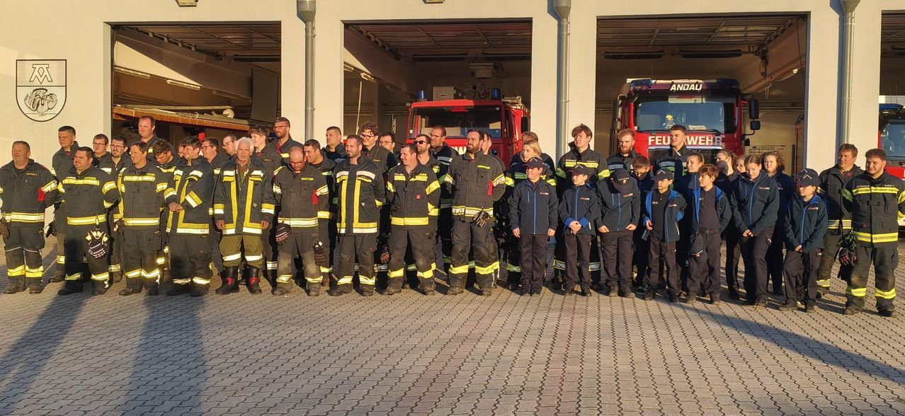 Eine Gruppe Feuerwehrleute und Kinder stehen vor Feuerwehrwagen auf einem Feuerwehrhaus.
