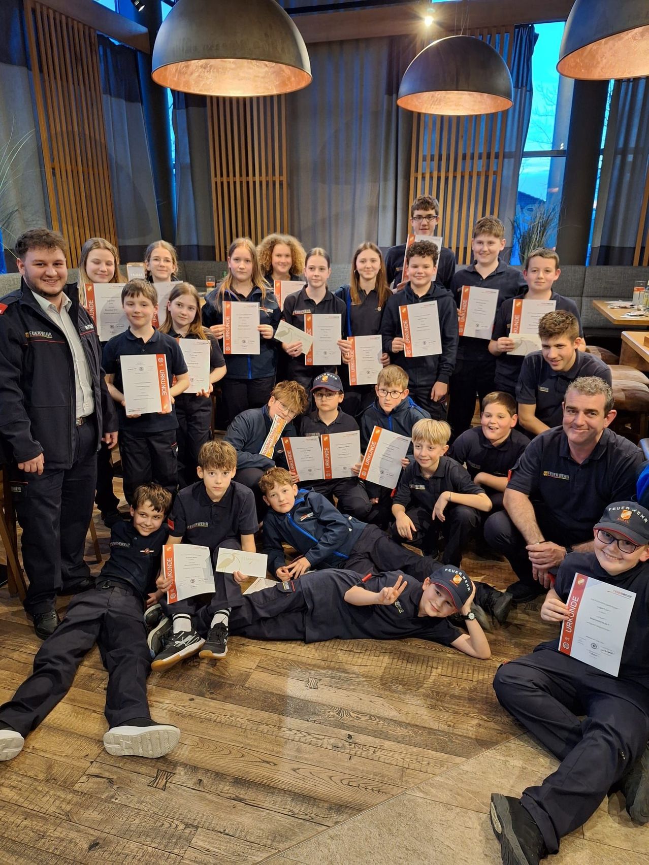 Gruppe junger Menschen und Erwachsener, die mit Zertifikaten in einem Hotel posieren. Die Zertifikate sind orange und weiß. Sie tragen einheitliche Kleidung.