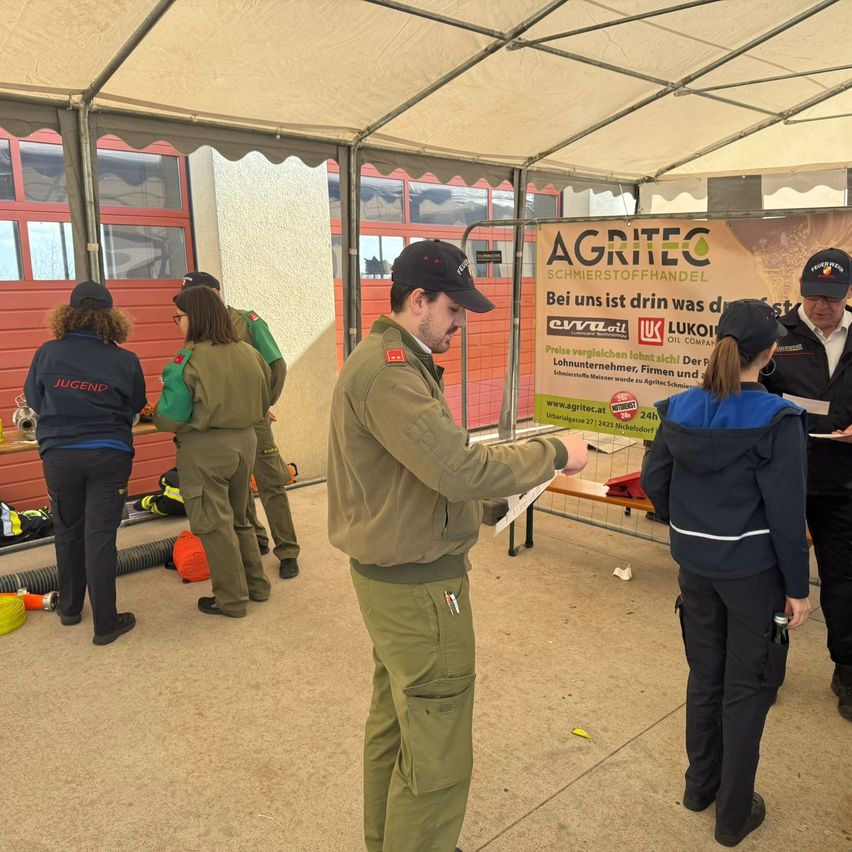 Eine Gruppe von Menschen in Uniformen steht in einem Zelt. Ein Mann spricht mit einer Frau und hält ein Stück Papier in der Hand. In der Nähe ist ein Banner sichtbar.
