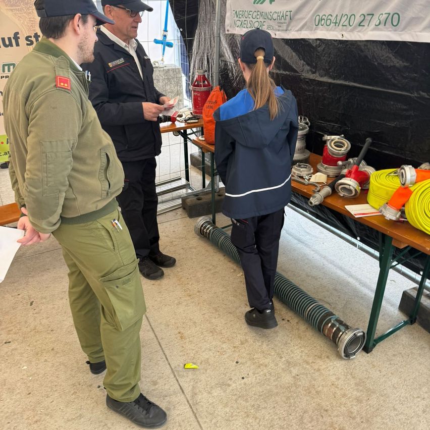 Drei Personen stehen um einen Tisch mit diversen Ausrüstungsgegenständen, darunter ein Schlauch, Werkzeuge und ein orangefarbener Behälter. Im Hintergrund ist ein Banner mit einer Telefonnummer zu sehen.