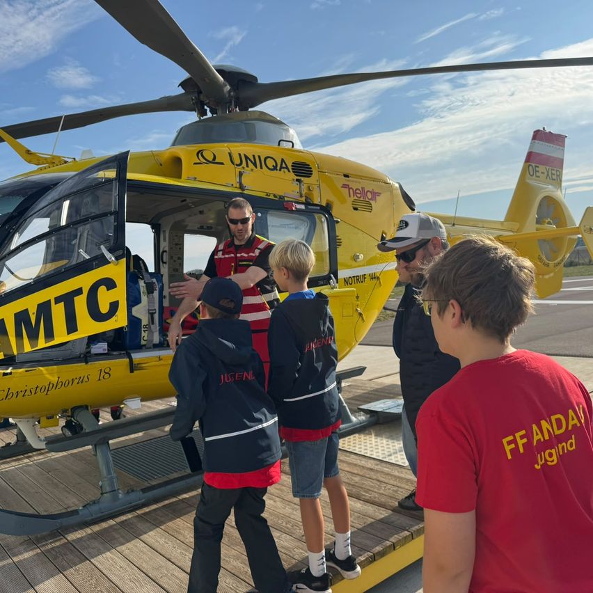 Eine Gruppe von Kindern und Erwachsenen steht um einen gelben Hubschrauber mit der Aufschrift 'MTC' herum. Einer der Erwachsenen trägt eine rote Weste und eine Sonnenbrille.