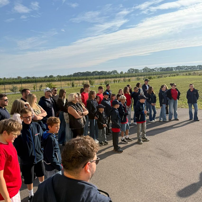 Eine Gruppe von Menschen, einschließlich Kindern, steht in einer Reihe auf einer Straße, mit einem Grasfeld und Bäumen im Hintergrund unter einem blauen Himmel.