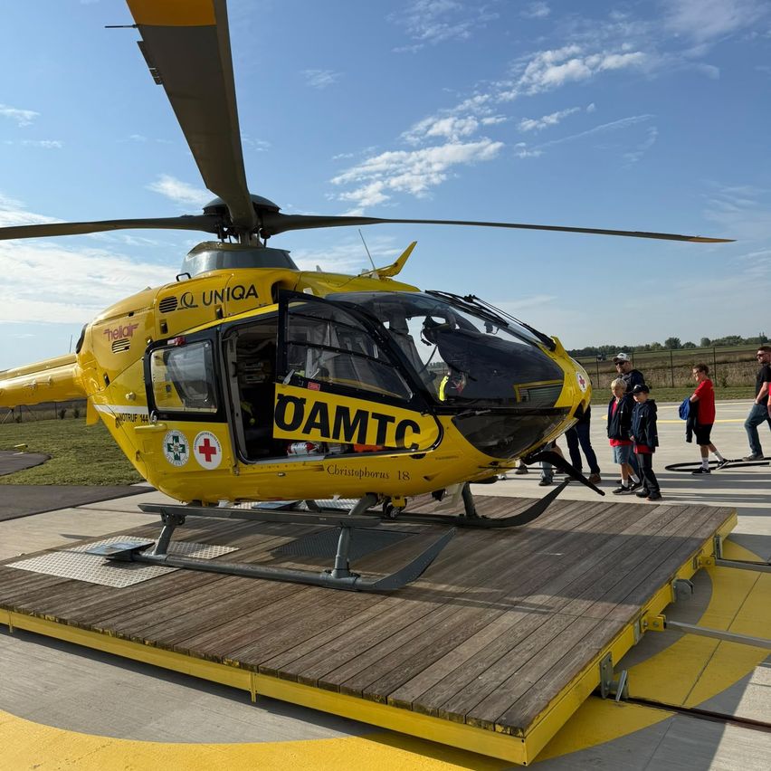 Ein gelber Hubschrauber mit der Aufschrift 'OAMTC' auf der Seite steht auf einer hölzernen Plattform. Mehrere Menschen stehen darum, darunter Kinder. Der Himmel ist blau mit einigen Wolken.