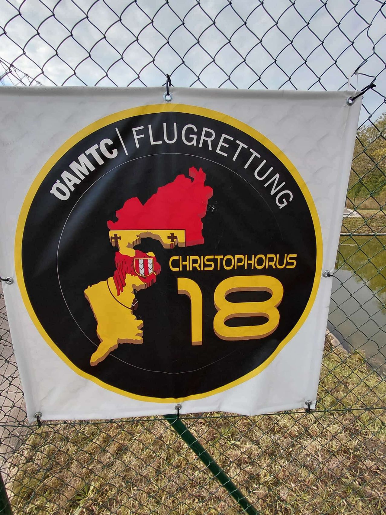Ein weißes Banner mit einem schwarzen Kreis in der Mitte, der eine Karte einer Region mit einem roten und gelben Design zeigt, die Worte 'OAMTC FLUGRETTUNG' über der Karte und 'CHRISTOPHORUS 18' darunter. Das Banner hängt an einem Zaun.