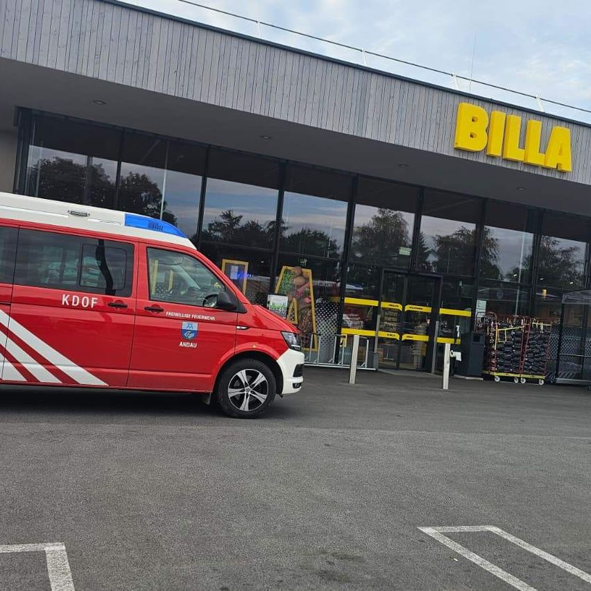 A red and white emergency vehicle is parked in front of the BILLA supermarket. The sky is clear and blue.