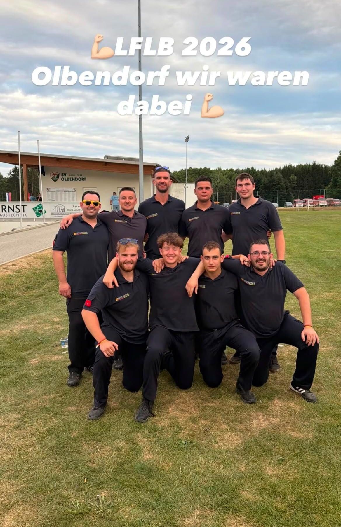 A group of men wearing black shirts and pants poses for a photo on a grassy field with a building and trees in the background.
