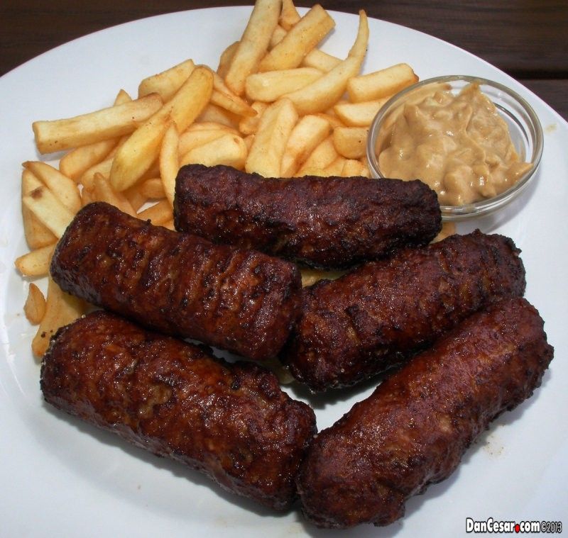 Ein weißer Teller mit gegrillten Würstchen und Pommes Frites. Der Teller hat auch eine kleine Schüssel mit cremiger Dipsauce.