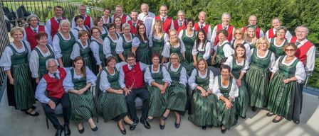 Eine Gruppe von Menschen in grüner und weißer traditioneller Kleidung posiert für ein Foto mit einer malerischen Aussicht auf Berge, einen See und eine Stadt im Hintergrund.
