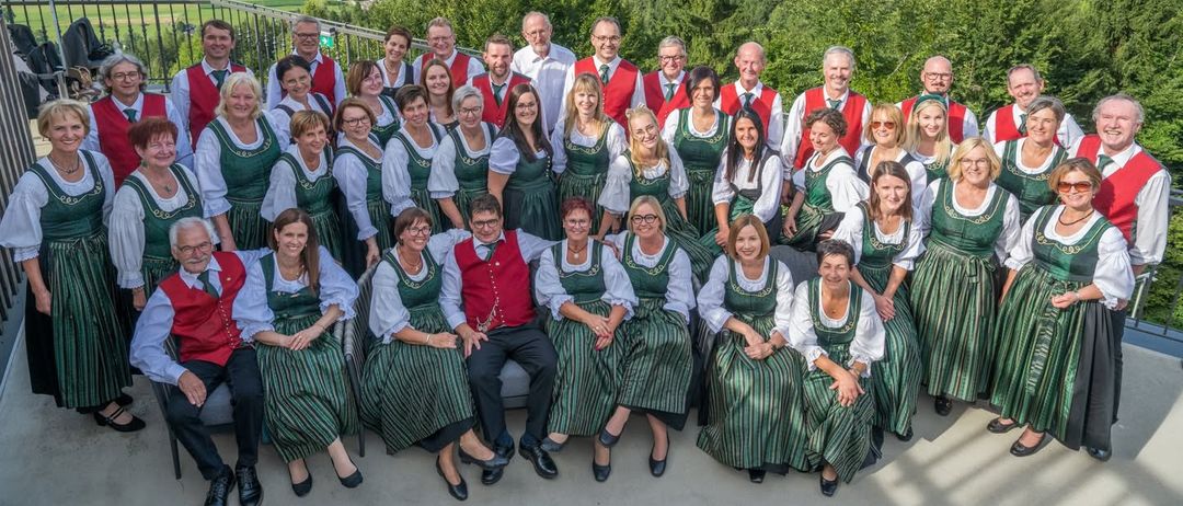 Eine Gruppe von Menschen in grüner und weißer traditioneller Kleidung posiert für ein Foto mit einer malerischen Aussicht auf Berge, einen See und eine Stadt im Hintergrund.