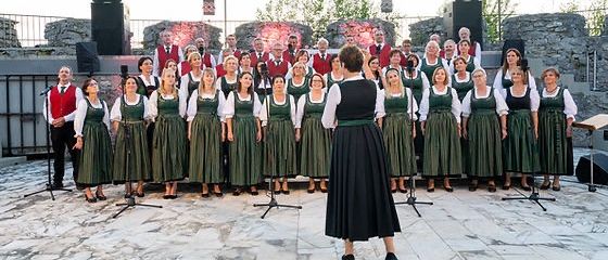 Ein Chor von Menschen in traditioneller Kleidung tritt auf einer Bühne mit Mikrofonen und Lautsprechern vor einer Steinmauer auf.