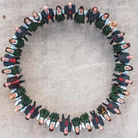 Eine Gruppe von Erwachsenen und Kindern in traditioneller Kleidung steht in einem Kreis auf einem Betonboden, möglicherweise für ein Foto.
