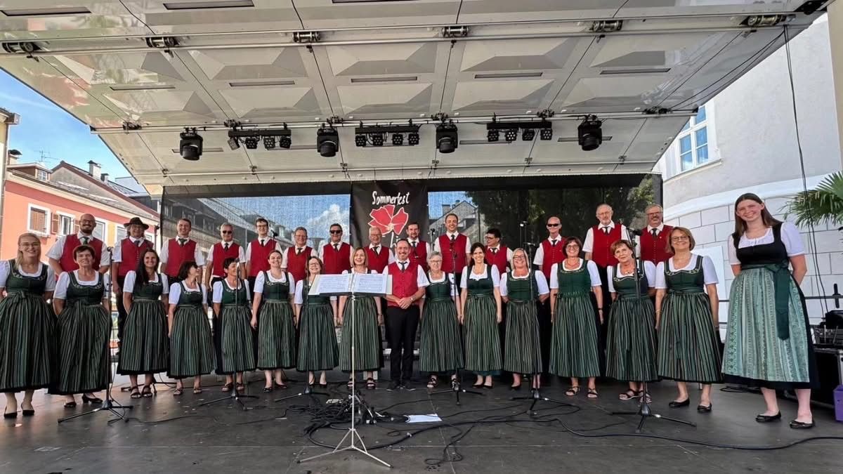 Ein Chor in traditioneller grüner und weißer Kleidung tritt auf der Bühne mit Notenblättern auf, vor einem Hintergrund mit einer roten Blume und einem Banner.